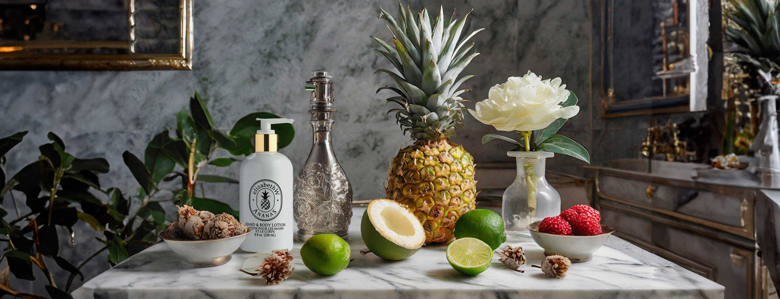 A marble countertop displays a variety of items including a pineapple, a halved coconut, limes, raspberries, lychees, a decorative bottle, a white lotion bottle with a pump, and a white flower in a small vase.