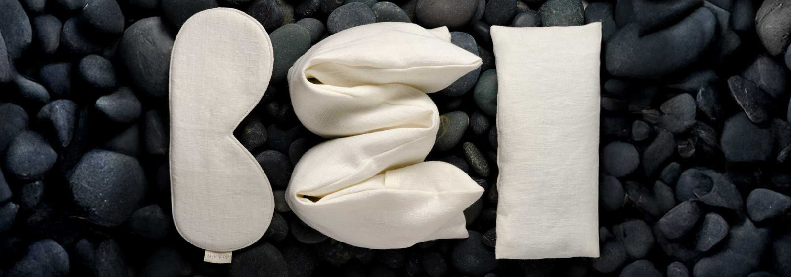 A cream-colored sleep mask, twisted fabric, and a rectangular pillow are arranged on smooth black stones, resembling the letters B, E, and D.