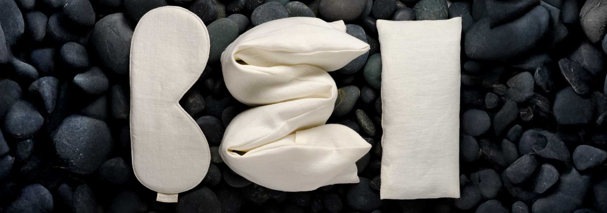 A cream-colored sleep mask, twisted fabric, and a rectangular pillow are arranged on smooth black stones, resembling the letters B, E, and D.