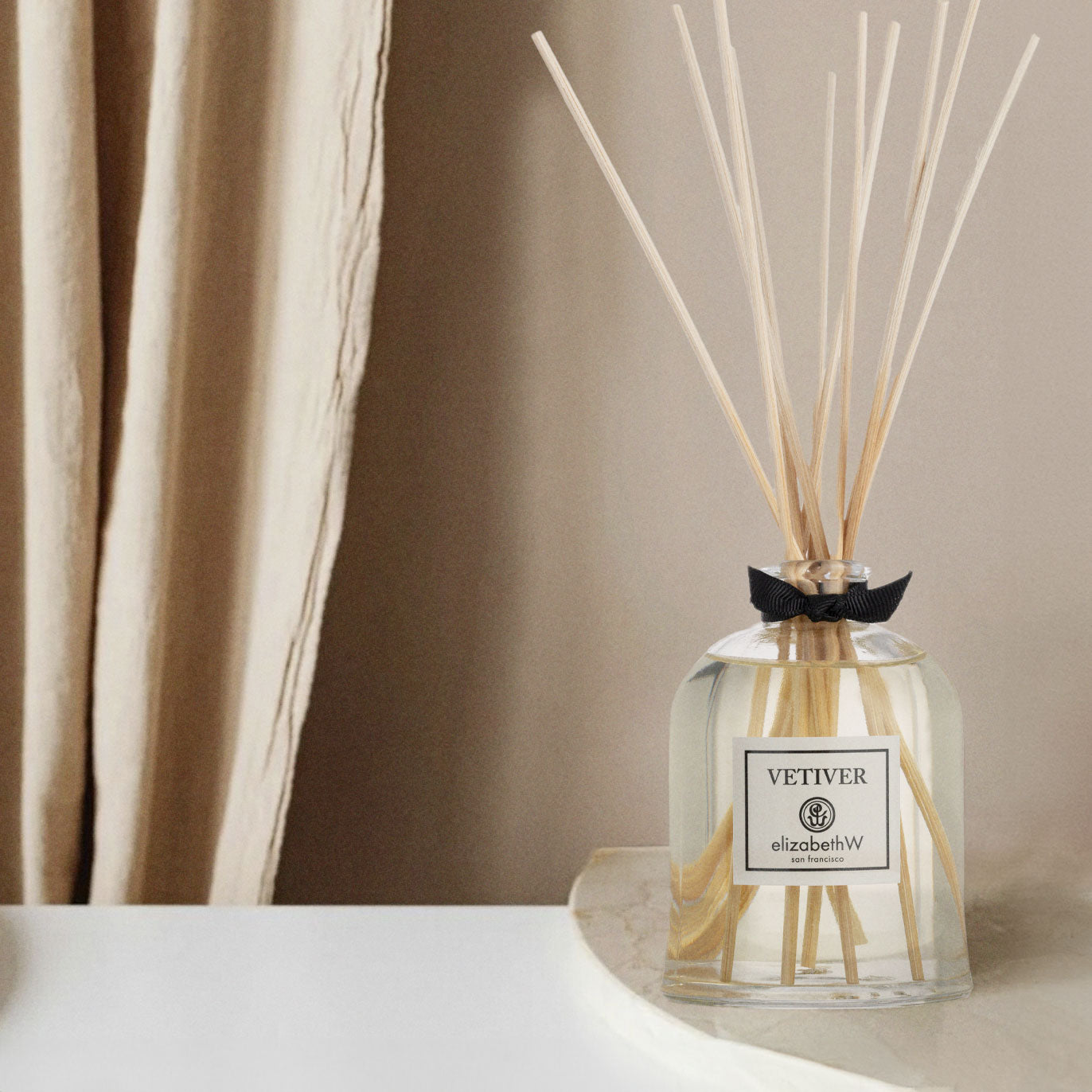 A reed diffuser with light-colored sticks in a clear glass bottle labeled "Vetiver elizabethW" is placed on a white marble surface. The background features a beige curtain.
