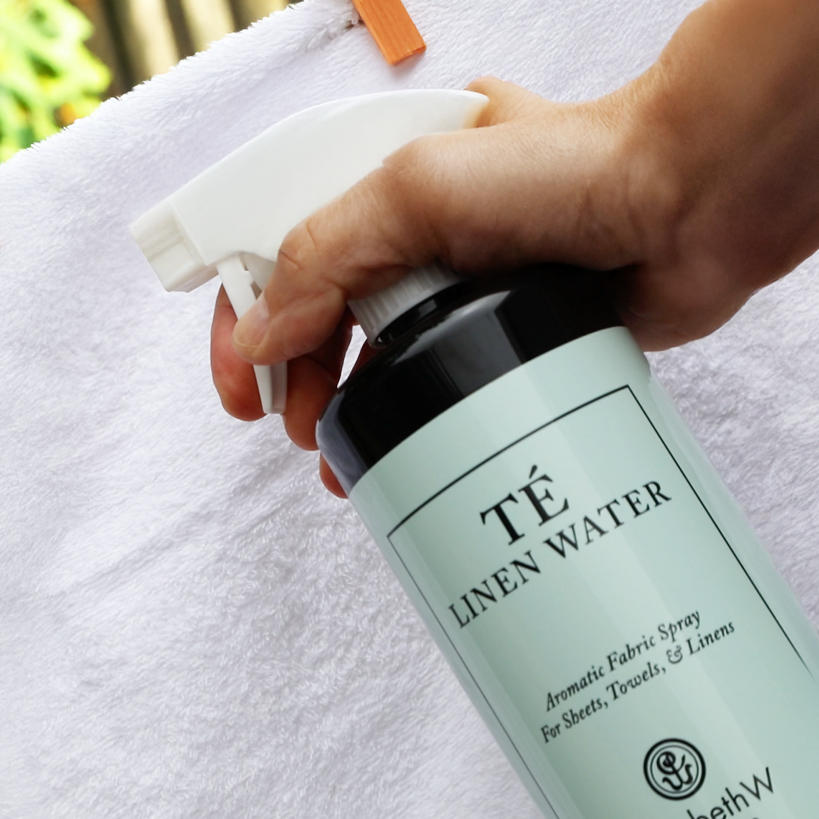 A hand holds a bottle labeled "TÉ Linen Water" and sprays it onto a white towel hanging on a clothesline with an orange clothespin.