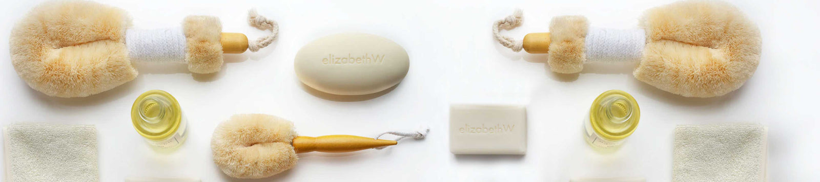 A set of natural skincare items including a dry brushing body brush, a soap bar, a small bottle of oil, a soft sponge, and a facial brush, all laid out on a white surface.
