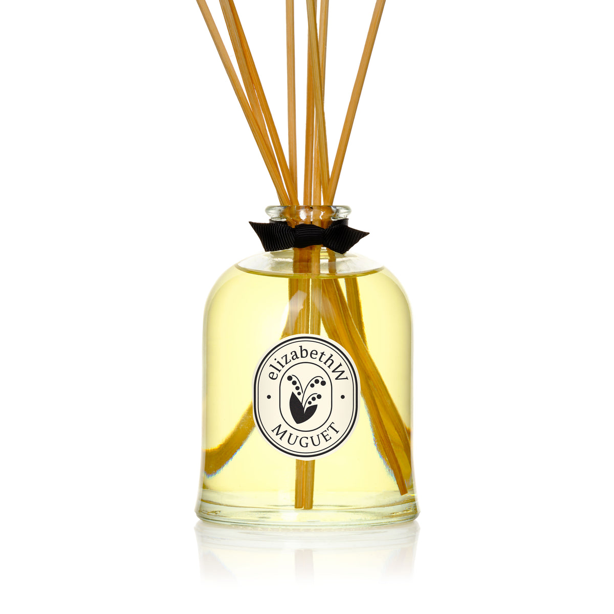 A glass bottle filled with yellow liquid and reed sticks, adorned with a black bow and a round "elizabethW Muguet" label—a stylish, eco-friendly diffuser from elizabethW offering long-lasting fragrance.