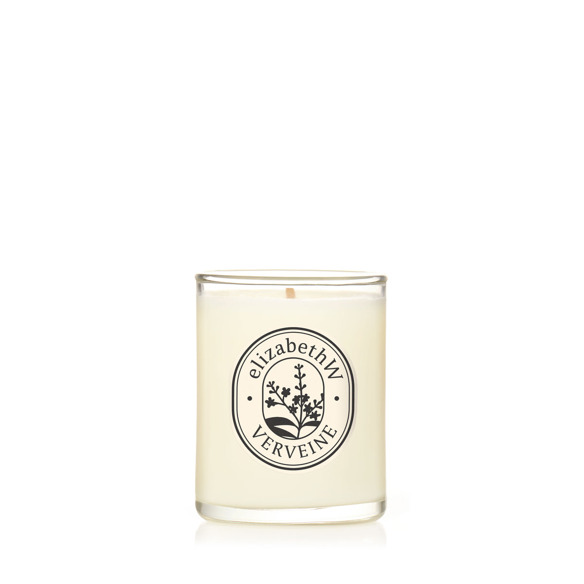A hand-poured white candle by elizabethW, named "Verveine — Petite Perfume Candle," sits in a clear glass holder with a botanical illustration and label on the front, all set against a plain white background.