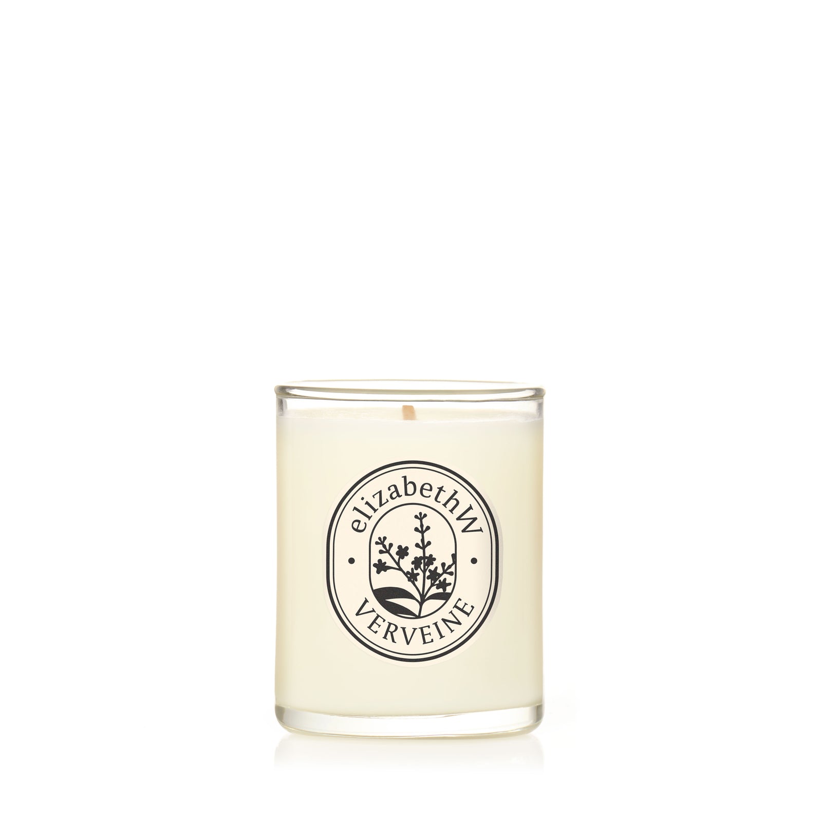 A hand-poured white candle by elizabethW, named "Verveine — Petite Perfume Candle," sits in a clear glass holder with a botanical illustration and label on the front, all set against a plain white background.